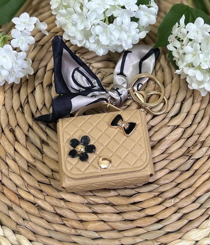 May include: A small, tan faux leather handbag keychain with a black and gold flower, a black bow, and a gold chain with a key ring. The keychain is on a woven straw surface.