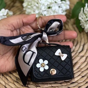 May include: Black quilted keychain wallet with a white bow, flower, and a black and white patterned scarf attached.