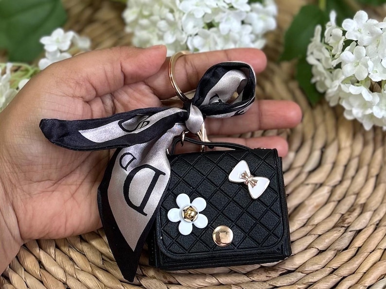 May include: Black quilted keychain wallet with a white bow, flower, and a black and white patterned scarf attached.