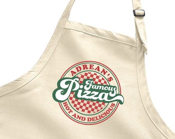 Op de afbeelding: Een beige keukenschort met een grote rode en witte geruite cirkel met de tekst "Adrean's Famous Pizza Hot and Delicious" in groen.