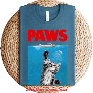 K&ouml;nnte beinhalten: T-Shirt in Heather Deep Teal mit dem Wort "PAWS" in roten Buchstaben. Das Design ist eine Parodie auf den Film "Jaws" und zeigt eine Katze, die aus dem Wasser greift. Das T-Shirt liegt auf einer gewebten Oberfl&auml;che.