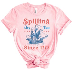 May include: A pale pink t-shirt with the words "Spilling the Tea Since 1773" in red and blue. A blue illustration of a sailing ship is centered on the shirt, with stars on either side.