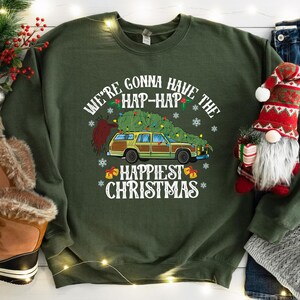 May include: Dark green sweatshirt with the text "WE'RE GONNA HAVE THE HAP-HAP HAPPIEST CHRISTMAS" and a graphic of a station wagon carrying a Christmas tree. The design includes snowflakes and bells. A festive gnome and Christmas decorations are in the background.