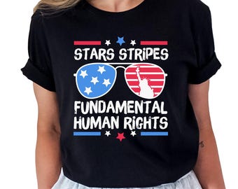 Puede incluir: Camiseta negra con un diseño patriótico. El gráfico presenta gafas de sol con estrellas azules y rayas rojas, la Estatua de la Libertad y el texto "STARS STRIPES FUNDAMENTAL HUMAN RIGHTS."