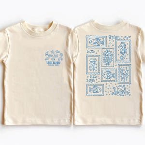 May include: Cream-colored t-shirt with blue sea-themed graphics. The front features a cluster of sea creatures and text that reads "WHITE FAMILY Summer 2026." The back displays various sea creatures in stamp-like designs.