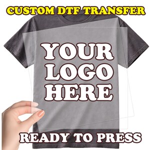 May include: A gray t-shirt with a clear transfer sheet featuring the text "YOUR LOGO HERE" in a bold, white font with a brown outline. The words "CUSTOM DTF TRANSFER" and "READY TO PRESS" are also visible.