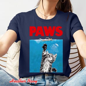 K&ouml;nnte beinhalten: Marineblaues T-Shirt mit einem Parodie-Grafik. Die Grafik zeigt das Wort "PAWS" in roten Blockbuchstaben &uuml;ber einem blauen Rechteck. Im Rechteck greift eine Katze nach einem Schwimmer im Wasser. Das Shirt ist aus weichem Material.
