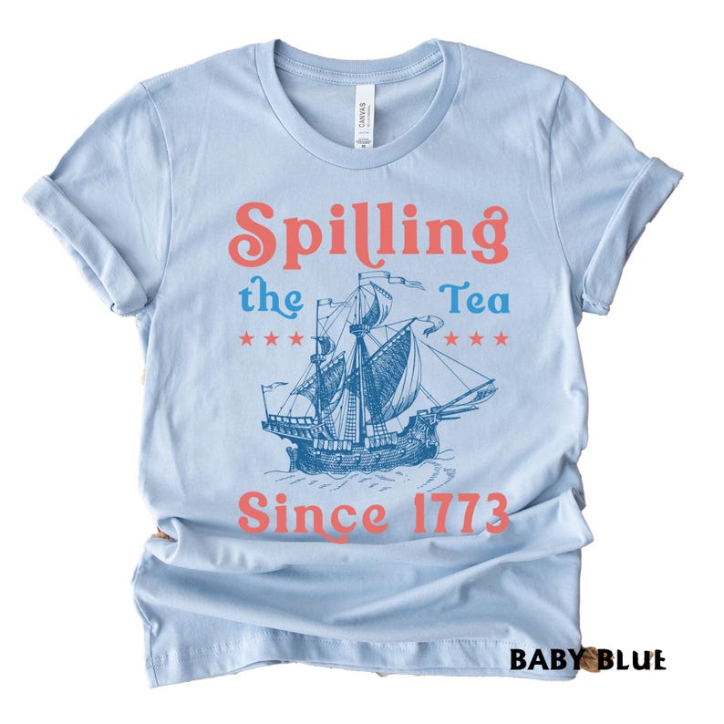 May include: Pale blue t-shirt with rolled sleeves. The shirt displays a ship graphic and the text "Spilling the Tea Since 1773" in coral and blue. The shirt is made of a soft material.