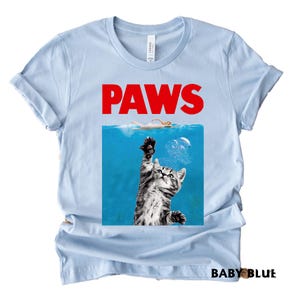 K&ouml;nnte beinhalten: Hellblaues T-Shirt mit dem Wort "PAWS" in roter Schrift. Das Design ist eine Parodie auf ein Filmplakat, mit einer Katze, die aus dem Wasser nach einem Schwimmer greift. Die Worte "BABY BLUE" sind unten aufgedruckt.