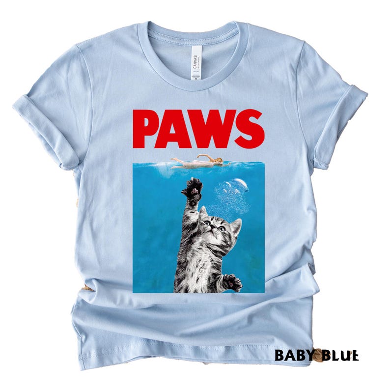 K&ouml;nnte beinhalten: Hellblaues T-Shirt mit dem Wort "PAWS" in roter Schrift. Das Design ist eine Parodie auf ein Filmplakat, mit einer Katze, die aus dem Wasser nach einem Schwimmer greift. Die Worte "BABY BLUE" sind unten aufgedruckt.