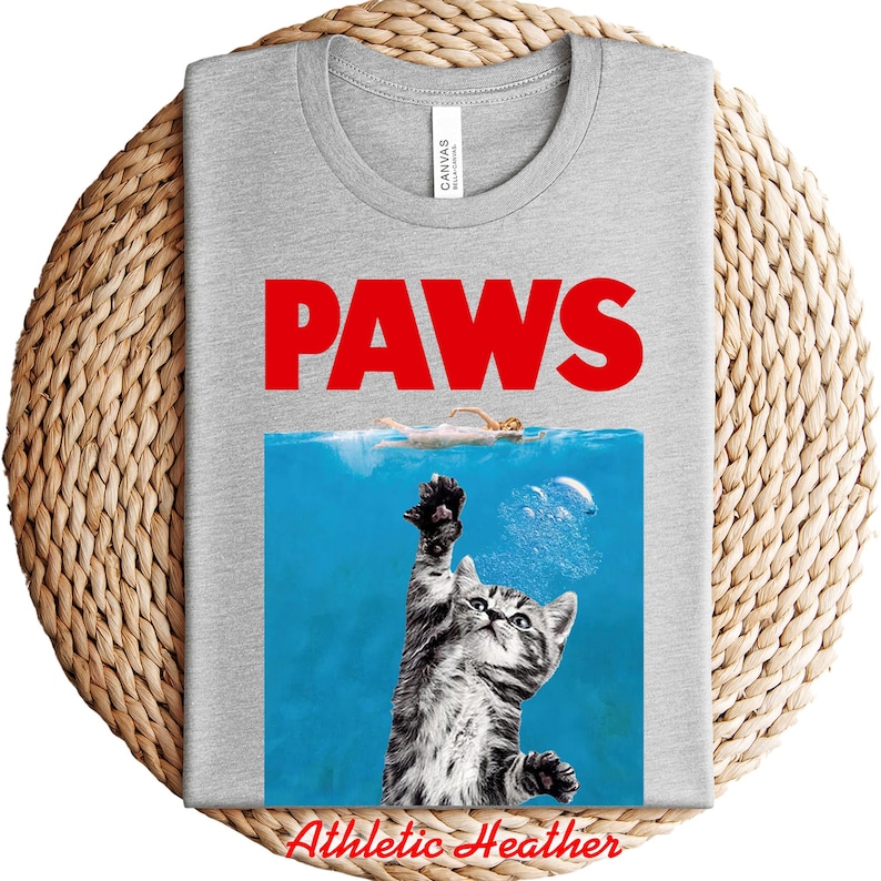 K&ouml;nnte beinhalten: Hellgraues T-Shirt mit einem Parodie-Design. Das Design zeigt das Wort "PAWS" in Rot, eine Katze, die aus dem Wasser greift, und eine Schwimmerin. Das Shirt besteht aus einem weichen, bequemen Material. Der Text "Athletic Heather" steht unten.