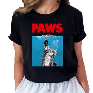 K&ouml;nnte beinhalten: Schwarzes T-Shirt mit dem roten Schriftzug "PAWS". Das Design zeigt eine grau-wei&szlig;e Katze, die aus blauem Wasser nach oben greift, mit einer Person, die dar&uuml;ber schwimmt.