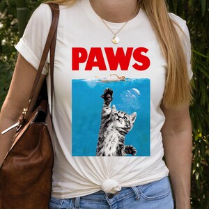 K&ouml;nnte beinhalten: Wei&szlig;es T-Shirt mit dem Wort "PAWS" in roten Blockbuchstaben. Das Shirt zeigt eine Grafik einer Katze, die aus blauem Wasser nach einem Schwimmer greift. Die Katze ist schwarz und grau gestreift.
