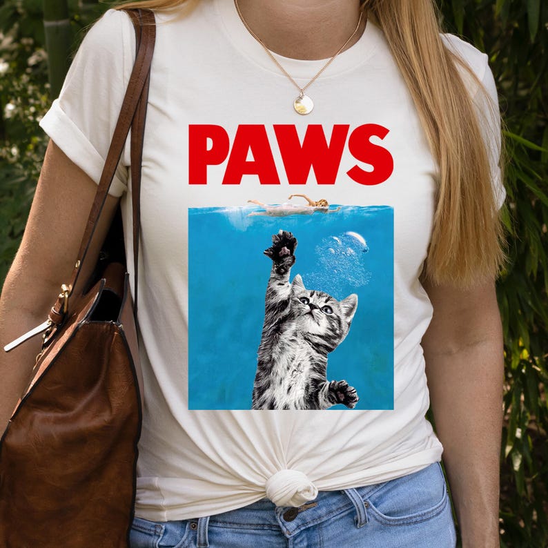 K&ouml;nnte beinhalten: Wei&szlig;es T-Shirt mit dem Wort "PAWS" in roten Blockbuchstaben. Das Shirt zeigt eine Grafik einer Katze, die aus blauem Wasser nach einem Schwimmer greift. Die Katze ist schwarz und grau gestreift.