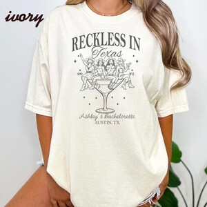 May include: Ivory-colored t-shirt with the text "RECKLESS IN Texas" and a graphic of women in cowboy hats in a martini glass. The text "Ashley's Bachelorette, Austin, TX" is also printed on the shirt.