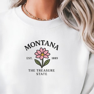 May include: White sweatshirt featuring the word "MONTANA" above a pink flower graphic with green leaves. Below the flower, the text reads "EST. 1889" and "THE TREASURE STATE". The sweatshirt has a crew neck and long sleeves.