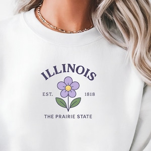 Op de afbeelding: Witte sweatshirt met het woord "ILLINOIS" in paarse boogtekst boven een paarse bloem met een gele kern. Daaronder staan de woorden "EST. 1818" en "THE PRAIRIE STATE". Een casual kledingstuk met staatsthema.
