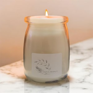 Puede incluir: Una vela blanca en un tarro de vidrio transparente con una etiqueta blanca que dice "simplistik Beauty Co." y un simple dibujo de línea negra de una rama con hojas.