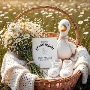 May include: A wicker basket filled with a bouquet of white daisies, a white plush duck, a card that reads "Here we grow again" and a pair of white crochet baby booties. The basket is lined with a cream-colored crocheted blanket.