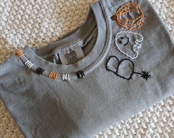 May include: A grey crewneck t-shirt with a Comfort Colors label. The neckline features orange, white, and black embroidered stitching. The front displays an embroidered orange pumpkin, a white ghost, and a black spider with a web-like line.