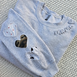 Op de afbeelding: Een lichtgrijs sweatshirt met het woord "mama" geborduurd op de borst. De linkermouw heeft drie hartvormige patches: een met een rode bloemenprint, een met camouflage en een met een zwart-witte hondenprint.