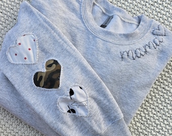 Könnte beinhalten: Ein hellgraues Sweatshirt mit dem Wort "Mama" auf der Brust gestickt. Der linke Ärmel hat drei herzförmige Aufnäher: einer mit rotem Blumendruck, einer mit Tarnmuster und einer mit einem schwarz-weißen Hundeaufdruck.