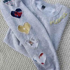 May include: A pale grey sweatshirt with yellow embroidered "mama" lettering. The left sleeve has five heart-shaped patches with diverse patterns, including stripes, a fire engine, and a sun. The sweatshirt is made of a soft fabric.