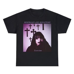 Könnte beinhalten: Schwarzes T-Shirt mit einem Grafikdesign. Das Design zeigt den Text "SIDEWALKS AND SKELETONS" über einem Bild einer Person mit Kreuzen und dem Wort "Exorcism". Das Bild hat einen lila und weißen Hintergrund.