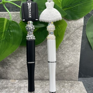 May include: Two pens with decorative tops shaped like a bride and groom. The bride is wearing a white dress and the groom is wearing a black tuxedo. The pens are both black and white.