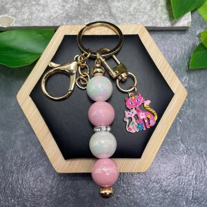 May include: A gold-toned metal keychain featuring a central vertical row of four alternating iridescent white and solid pink spherical beads, separated by a small rhinestone spacer, with a pink and gold cat charm attached to a lobster clasp.