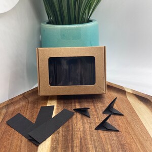 May include: A box of black origami paper with a window showing the paper inside. There are four black origami triangles and three black origami strips on a wooden surface.