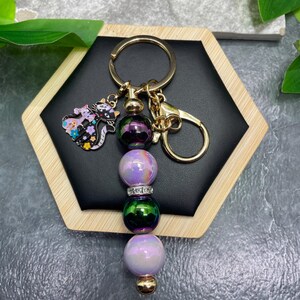 May include: A gold-toned metal keychain featuring a dangling charm of two cats decorated with colorful flowers. The keychain includes a series of four spherical beads in iridescent purple, dark green, and light purple, with a silver-toned rhinestone spacer.