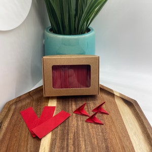 May include: A box of red origami paper with a few folded origami triangles and strips of red paper on a wooden tray.