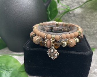 May include: A set of three bracelets with brown wooden beads, gold accents, and a crystal charm. The bracelets are stacked on top of each other and are displayed on a black velvet box.