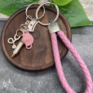 以下が含まれることがあります： ピンクの編み込みレザーキーホルダー。シルバーのキーリングと、ナースキャップ、フェイスマスク、注射器、ハサミなどのチャームが付いています。