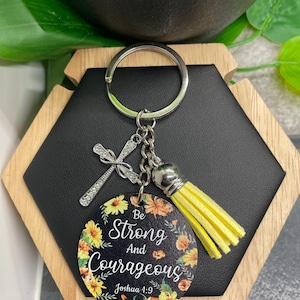 Puede incluir: Un llavero plateado con un dije de cruz y una borla amarilla. El colgante redondo presenta el texto "Be Strong And Courageous" rodeado de ilustraciones florales. El llavero está sujeto a un llavero plateado.