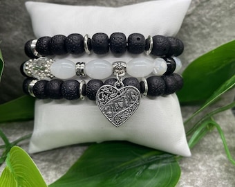May include: Three layered bracelets featuring black lava beads, white oval beads, and a silver heart charm with the word "Mom" engraved on it.