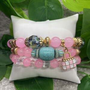 May include: A set of four pink and turquoise beaded bracelets with gold accents. The bracelets feature a variety of beads, including round, square, and rectangular shapes. One bracelet has a small pink and white handbag charm.