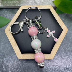 May include: A keychain featuring a silver-tone metal ring and a lobster clasp. The central design consists of three round, iridescent beads in shades of pink and white, separated by silver-tone rhinestone spacers. A small silver-tone cross charm with pink enamel accents hangs from the ring.
