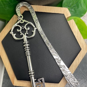 May include: Silver key-shaped bookmark with a floral design. The bookmark is attached to a decorative key with a silver finish.