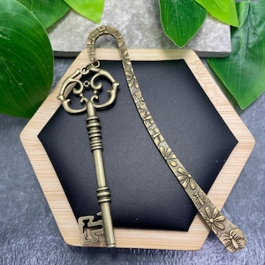 May include: Antique brass key and floral bookmark set on a black hexagon-shaped display. The key has ornate detailing, and the bookmark features a repeating flower pattern. The display has a wooden frame.