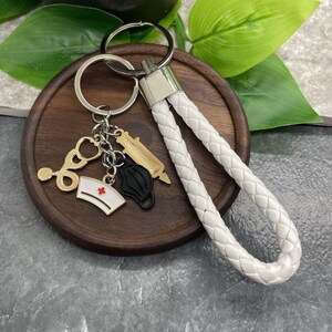 以下が含まれることがあります： 白い編み込み革のキーホルダーに、金と銀のチャームがセットになっています。チャームには、聴診器、注射器、看護師の帽子、フェイスマスクが含まれています。