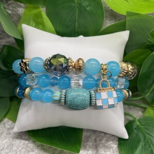 May include: A set of four bracelets featuring blue and turquoise beads, gold accents, and a small blue and white checkered charm in the shape of a handbag.