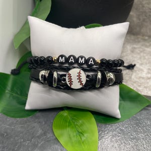 May include: A set of three bracelets with black beads, black leather, and silver accents. The middle bracelet features a white baseball charm with red stitching. The top bracelet has the word "MAMA" spelled out in black letters.