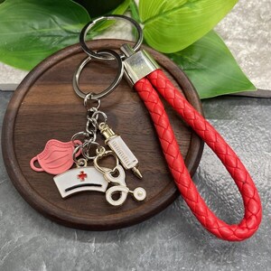 以下が含まれることがあります： 赤い編み込みキーホルダー。シルバーのキーリングと、赤い十字のついた白いナースキャップ、聴診器、注射器、ピンクのマスクなどのチャームが付いています。