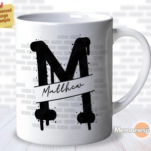 Puede incluir: Taza de cerámica blanca con una letra "M" negra de estilo graffiti y el nombre "Matthew" escrito en una pancarta debajo. La taza tiene un patrón de pared de ladrillo.