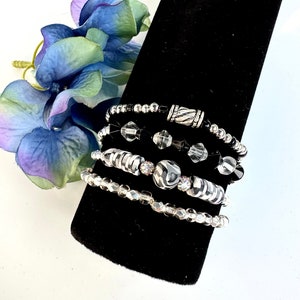 Può includere: Set di cinque braccialetti con perline nere e trasparenti e dettagli argentati. Alcuni braccialetti presentano un motivo zebrato. I braccialetti sono esposti su un supporto di velluto nero, con un fiore blu e viola sullo sfondo.