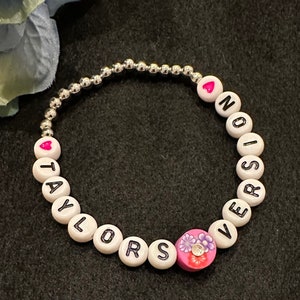 Könnte beinhalten: Ein Perlenarmband mit weißen Buchstabenperlen mit der Aufschrift "TAYLORS VERSION" und einer rosa Blumenperle. Das Armband hat auch silberne Perlen und herzförmige Akzente. Das Armband befindet sich auf einem dunklen Hintergrund.