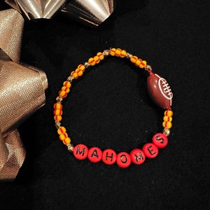 Könnte beinhalten: Ein Perlenarmband mit dem Namen "MAHOMES" in roten Buchstabenperlen. Das Armband hat gelbe und rote Perlen sowie einen footballförmigen Anhänger. Der Hintergrund ist schwarz.