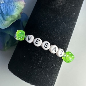 May include: A beaded bracelet spelling "VEGAS" with white letter beads. The bracelet features two translucent green dice beads and clear beads. The bracelet is displayed on a black velvet surface.